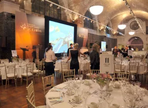 Banquet hall with arched brick ceiling and hanging lights, round tables set with white linens, glassware and numbered table signs, staff arranging chairs in front of a stage with large screens and orange award backdrops