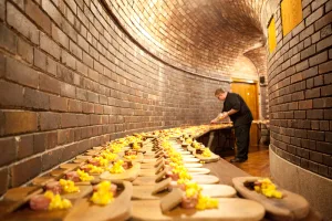 Chef arranging rows of wooden serving boards topped with small colourful canapés along a long curved brick‑walled corridor lit by warm lights.