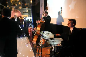 A jazz quartet in black suits performs at a formal event — drummer at a wooden drum kit in the foreground, an upright bassist plucking beside him with his shadow cast on a screen, and other musicians and tuxedoed guests blurred in the background.