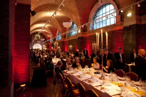 A formal dinner in a vaulted brick hall with arched windows, long white-clothed tables arranged with glassware, plates and tall candelabras, and guests in evening wear seated and mingling beneath warm red uplighting.