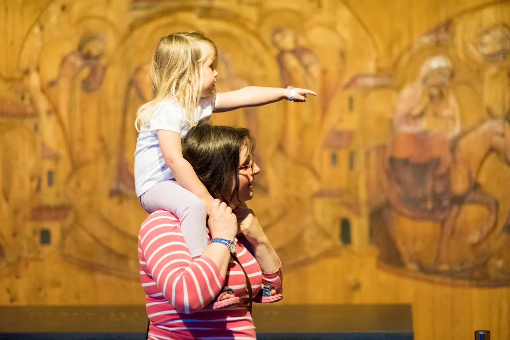 A woman in a pink-and-white striped top carries a young blonde girl on her shoulders while the girl points off to the side, set against a warm wooden wall with faded religious-style paintings.