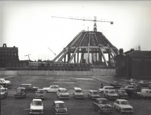 Black-and-white photograph of a large circular concrete structure under construction with cranes and exposed scaffolding, flanked by low buildings and a foreground dirt car park filled with vintage cars.
