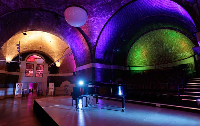 Grand piano on an empty stage beneath vaulted brick arches bathed in purple, green and amber lighting, with tiered seating and a large circular ceiling light in a dim performance venue.