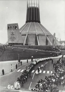 Modern circular concrete cathedral with a tall lantern tower looming over a long religious procession of robed clergy, uniformed band members and altar servers marching along a road while large crowds watch from the pavement.