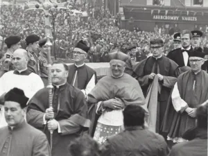 Black-and-white photo of a solemn clerical procession with robed priests and a bishop wearing a biretta at centre, a man carrying a tall ceremonial staff in front, and a large crowd with uniformed scouts and a shopfront reading ABERCROMBY VAULTS in the background.