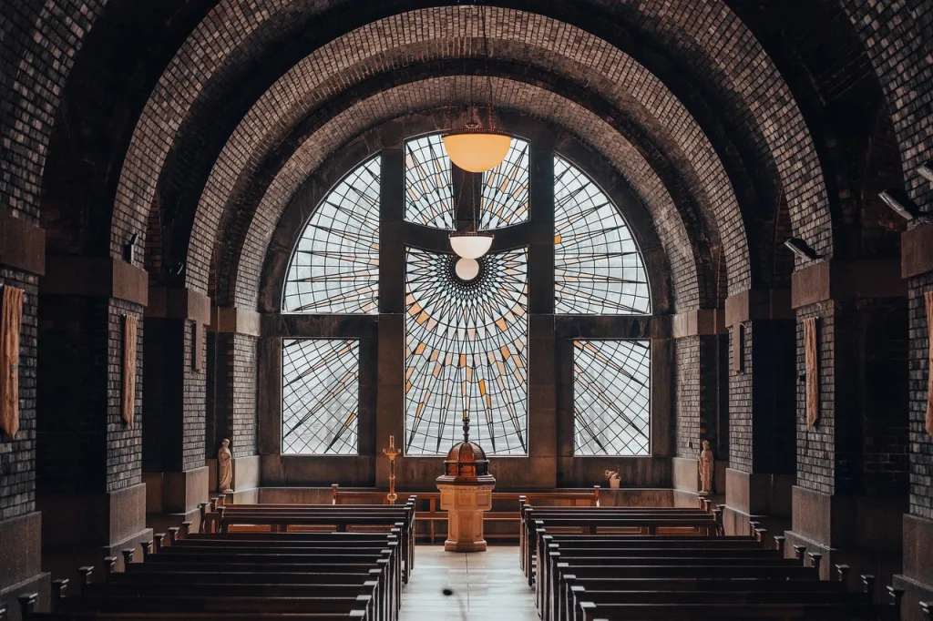 Symmetrical chapel interior with rows of wooden pews leading to a central baptismal font beneath a large arched stained-glass window with a sunburst radial pattern, framed by curved brick vaults and warm hanging lights.