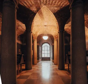 Symmetrical vaulted brick interior with thick stone columns flanking a central checkerboard-tiled aisle that leads to a large arched paned window, illuminated by a hanging globe lamp and warm amber light.