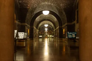 Long, dimly lit museum corridor with a series of brick barrel-vault arches and hanging globe lights, a glossy reflective floor, display panels and exhibits lining the sides, framed by large stone columns in the foreground.