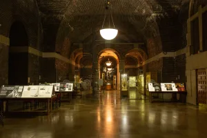 Dimly lit brick‑vaulted exhibition hall with arched ceilings, central hanging lamps and a spiral staircase, lined with illuminated display cases and information panels reflected on the polished floor.