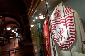 White liturgical vestment embroidered in red with wavy bands, a Chi‑Rho symbol and a crown motif, displayed behind glass in a dim, vaulted museum hall alongside other colourful robes.