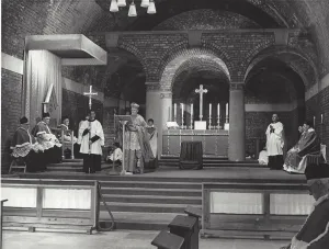 Clergy in ornate robes conduct a solemn Catholic service in a vaulted brick crypt, with a bishop reading at a lectern beside an altar lined with tall candles and a server holding a processional cross, seated priests at either side and wooden pews in the foreground.