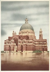 Large red‑brick cathedral with a prominent greenish dome topped by a cross, layered arched façades and a wide entrance reflected in calm water in the foreground beneath a pale, cloudy sky.