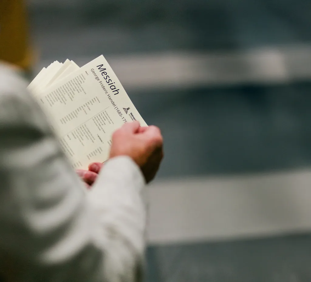 Hands and forearms of a person wearing a white jacket holding an open programme titled "Messiah" with printed song lists, set against a blurred auditorium background.