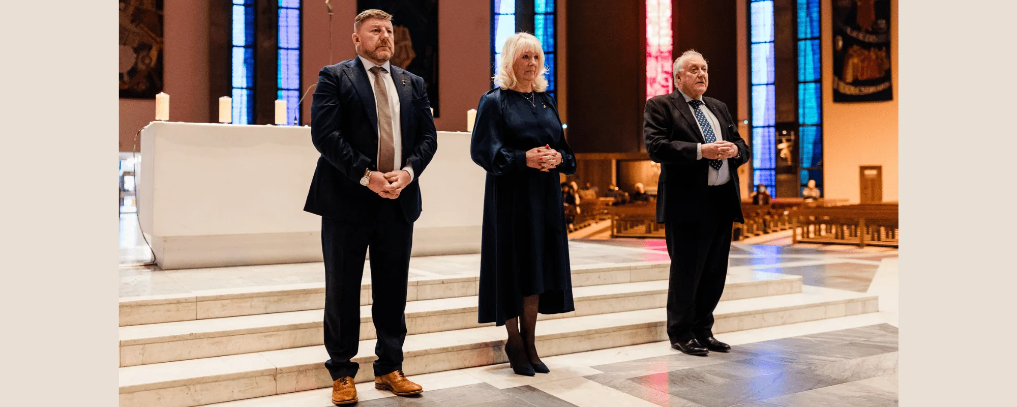 Three formally dressed adults—two men and a woman—stand solemnly on marble steps before a church altar with lit candles and tall stained-glass windows in the background.