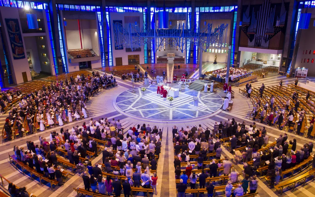 High-angle view of a modern circular cathedral interior showing a central altar where clergy in red robes lead a service beneath a suspended metal sculpture, surrounded by concentric wooden pews filled with worshippers, a patterned marble floor and tall blue stained-glass windows with organ pipes in the background.