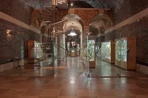 Vaulted brick gallery interior with arched ceilings, a central metal spiral staircase and glass display cases lining both sides illuminated with exhibits.