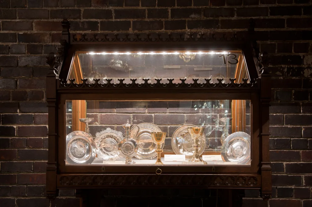 Ornate dark wooden glass display cabinet mounted on a brick wall, lit from above and showcasing reflective silver plates, goblets and ceremonial chalices with a small '13' visible in the upper right corner.