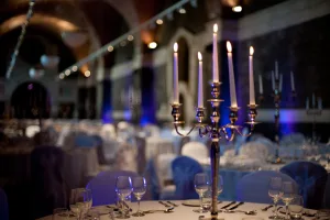 Silver five‑arm candelabrum with five lit white taper candles centred on a round, formally set banquet table with glassware and cutlery, blurred ballroom with chair covers and blue uplighting in the background.