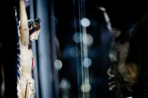 Close-up of a carved crucifix showing the figure of Jesus with a wound on his side, lit from the left against a dark background of blurred glass reflections and circular bokeh lights.