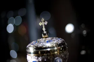 Close-up of an ornate silver lid or reliquary topped with a small cross on a spherical finial, decorated with blue enamel medallions and intricate filigree, set against a dark background with soft out-of-focus lights.