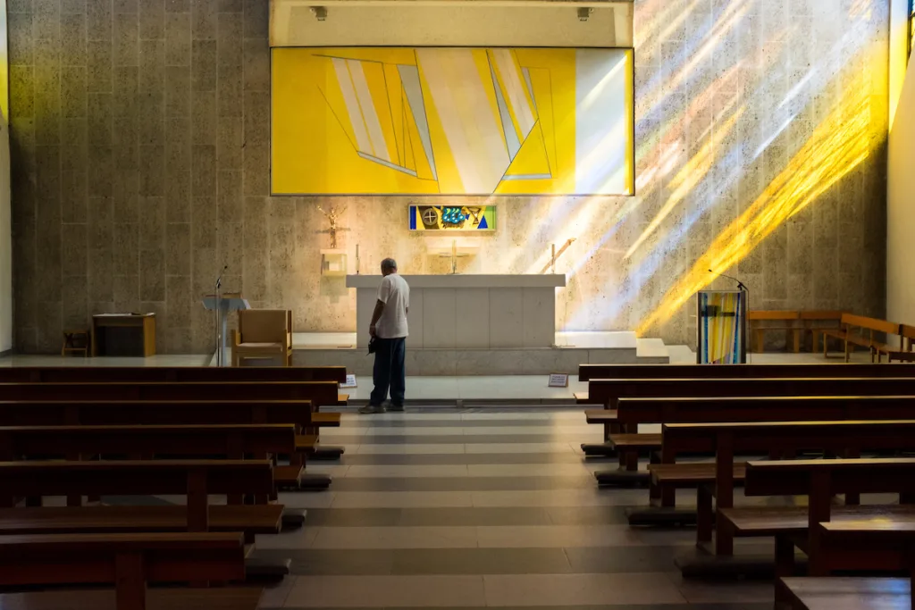 A man stands alone at the front of a modern church, facing a simple white altar beneath a large yellow abstract panel as sunlight from stained glass casts diagonal golden rays across the textured stone wall and rows of wooden pews.
