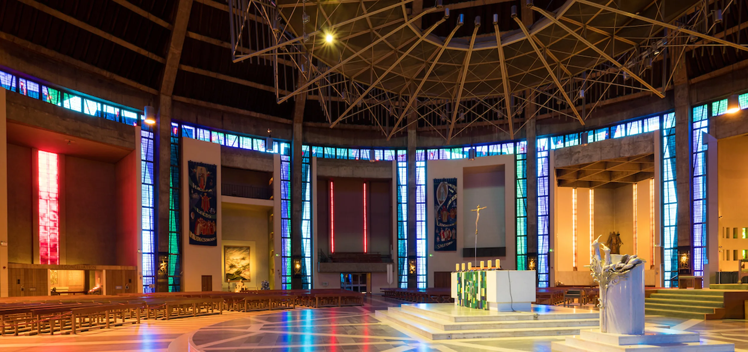 Spacious circular modern cathedral interior with a raised marble altar and crucifix at centre, colourful vertical stained-glass windows encircling the walls, a large geometric metal ceiling fixture above, rows of wooden benches and a sculpted lectern to the right.