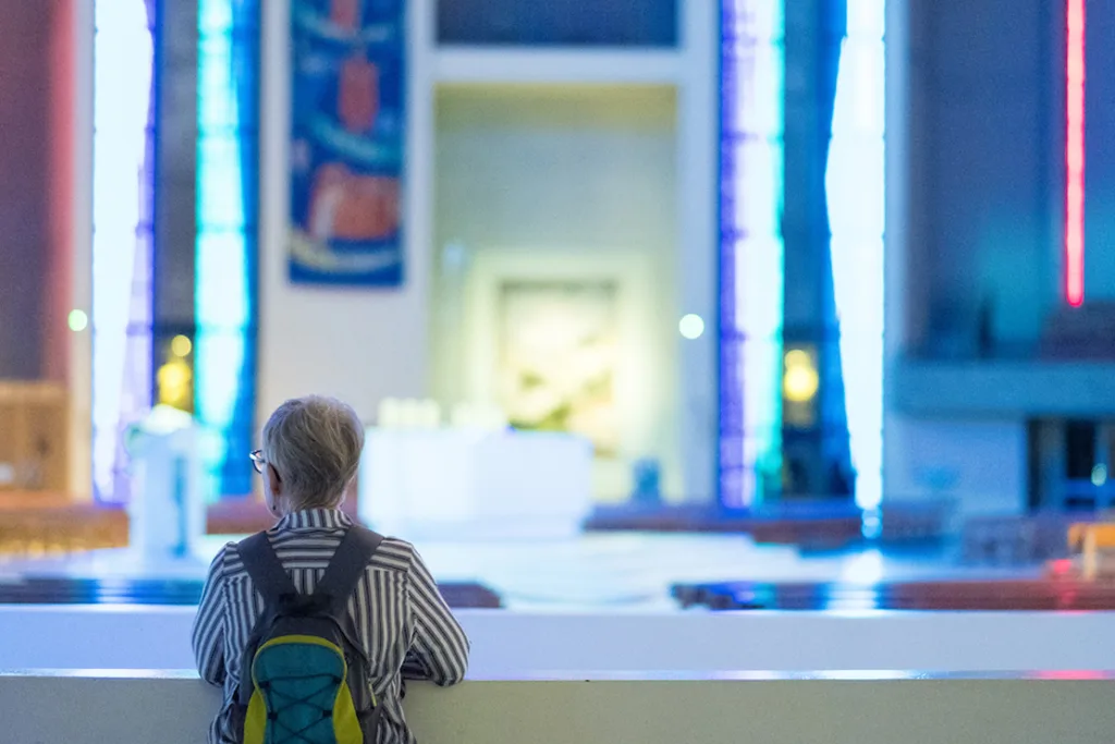 Older person seen from behind in a striped shirt with a small teal-and-yellow backpack, leaning on an interior ledge and looking toward a softly focused altar and tall vertical stained-glass windows lit in blue.