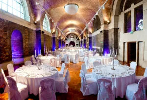 Long vaulted brick hall set for a formal banquet with round tables draped in white cloths and chair covers, candelabra centrepieces, parquet floor and purple uplighting beneath arched windows.