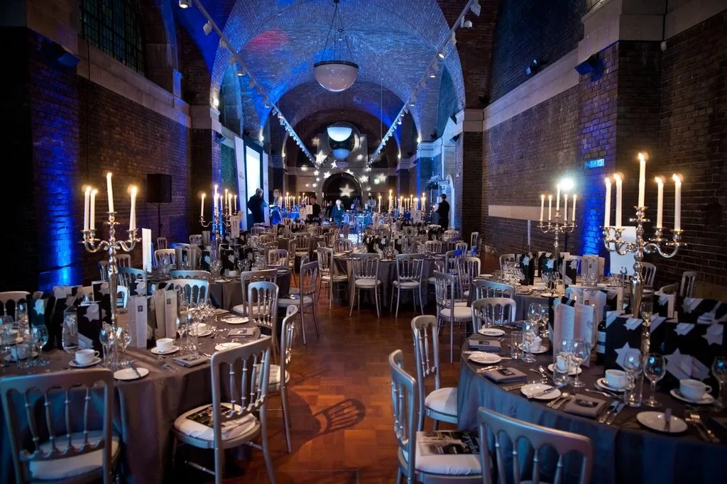 Long vaulted brick hall arranged for a formal dinner with rows of round tables draped in grey cloths, silver chairs, tall candelabra with lit candles and blue uplighting along the arched ceiling.