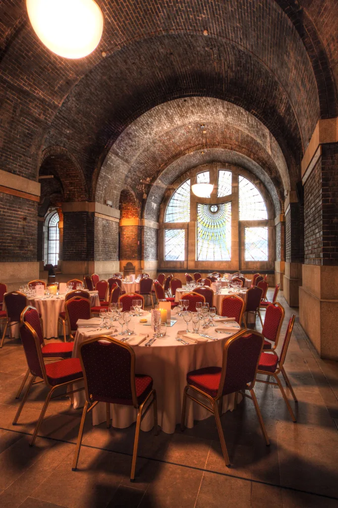 Vaulted brick-ceiling banquet hall with an arched stained-glass window, set with round tables draped in white linens, red-upholstered chairs and formal place settings with glassware and candles.