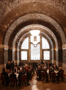 Formal dinner reception under a series of vaulted brick arches with a large decorative arched window and pendant chandeliers, guests in formal attire seated around round tables.