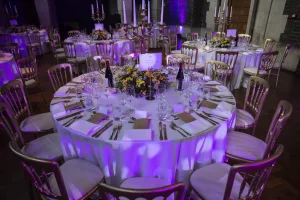 Round banquet table set for a formal dinner with a white tablecloth, gold chairs, a tall central candelabra and yellow floral centerpiece, wine bottles and multiple place settings with glassware under purple uplighting