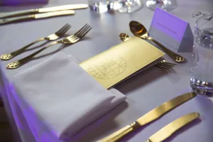 Formal place setting on a pale tablecloth featuring a folded white napkin, a gold menu card with a circular geometric design, polished gold cutlery, a folded place card and a water glass.