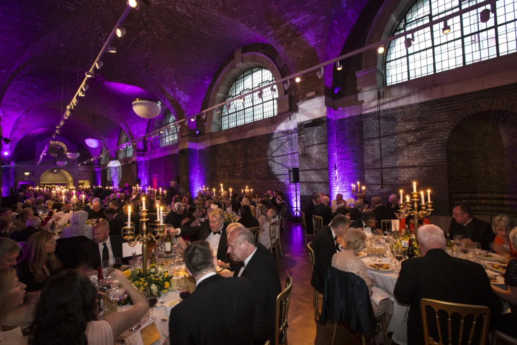 Formal black-tie banquet in a high vaulted brick hall with purple uplighting, rows of round tables set with candelabras and wine, and dozens of guests dining beneath large arched windows.