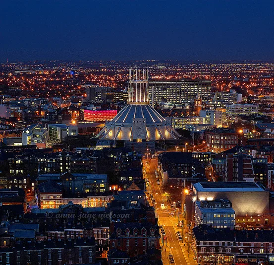 Night aerial view of a city centred on a modern circular cathedral with a conical, tent-like roof topped by a crown of vertical spires, surrounded by glowing streets and lit buildings.