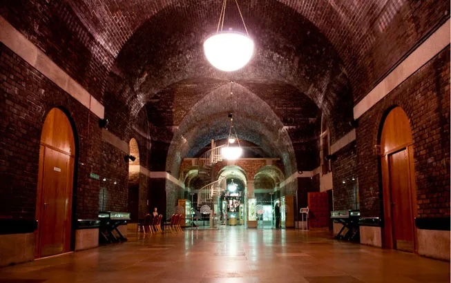 Spacious vaulted brick hall with dark arched ceilings, central hanging lights, a spiral staircase at the far end and wooden doors and scattered chairs along the sides.