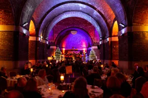 Wide vaulted brick hall lit in purple and orange, with a choir performing on a small stage flanked by two decorated, lit Christmas trees while a seated audience dines at candlelit tables.