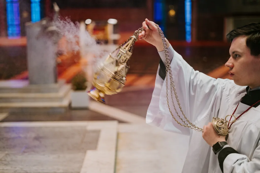A young altar server in a white surplice swings a smoking brass thurible by its chains inside a dim church, with blurred steps and stained-glass windows in the background.