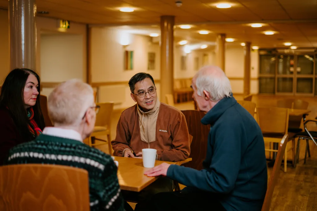 Four adults seated around a wooden table in a warmly lit community room, talking and smiling as a man wearing glasses leans forward with a white mug on the table.