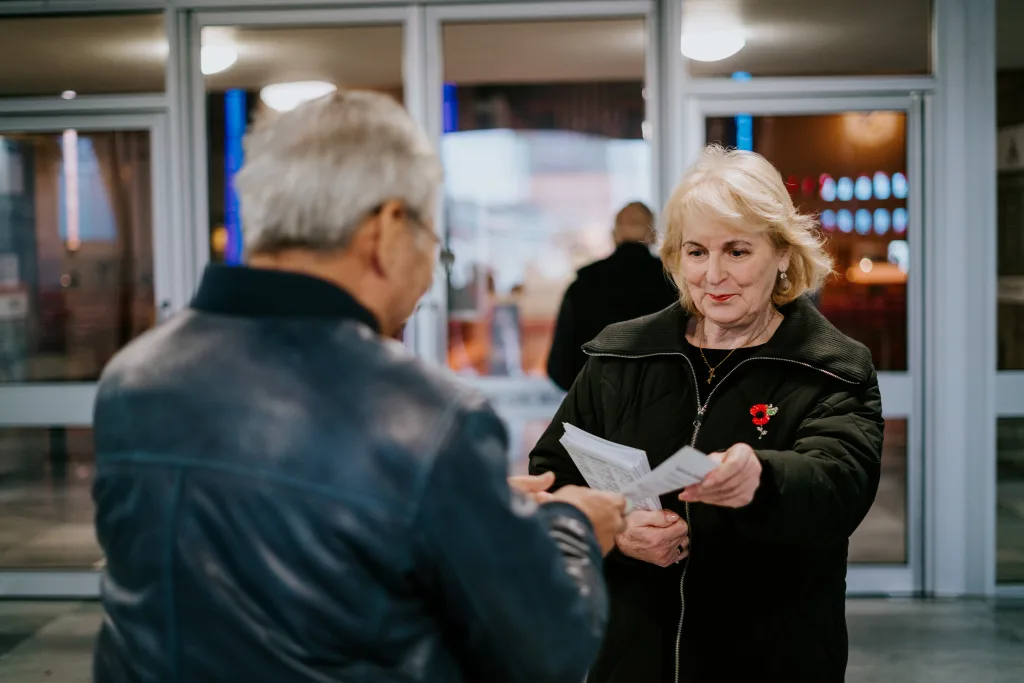 An older woman in a black coat wearing a red poppy pin hands papers to an older man near glass entrance doors, with blurred interior lights behind them.