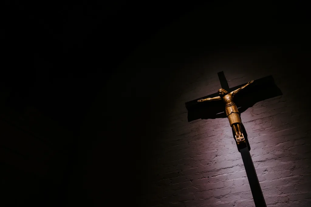 Gilded crucifix of Jesus mounted on a dark brick wall to the right of the frame, illuminated by a single overhead spotlight that casts a long shadow across the textured bricks.