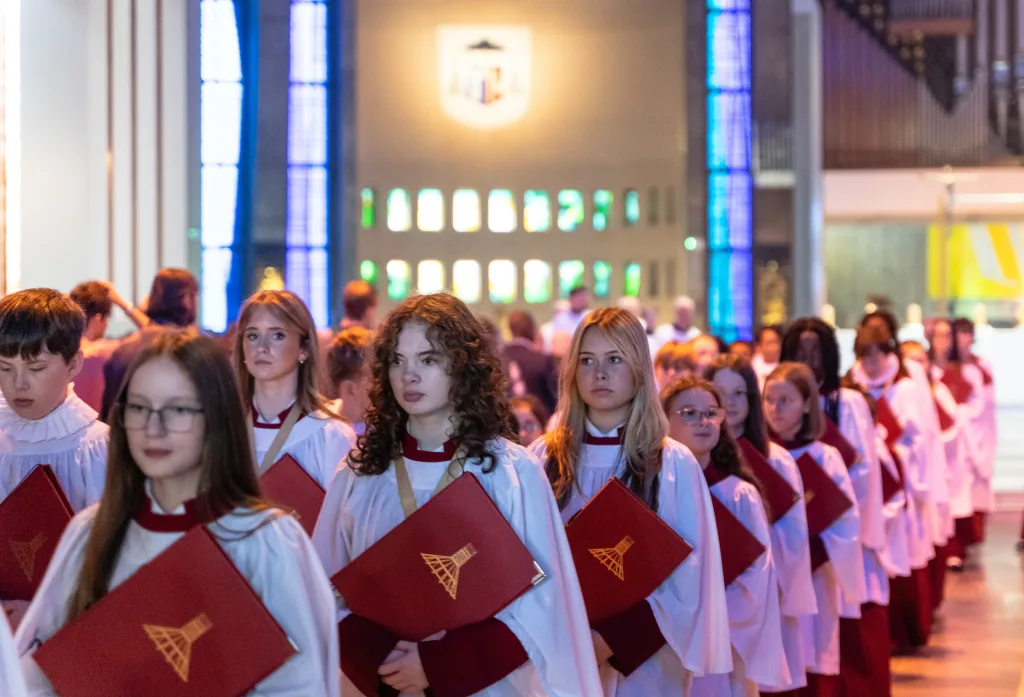 A line of young choristers in white robes with red collars holding red hymnals, processing down a church aisle with tall blue stained-glass windows and an illuminated altar in the background.