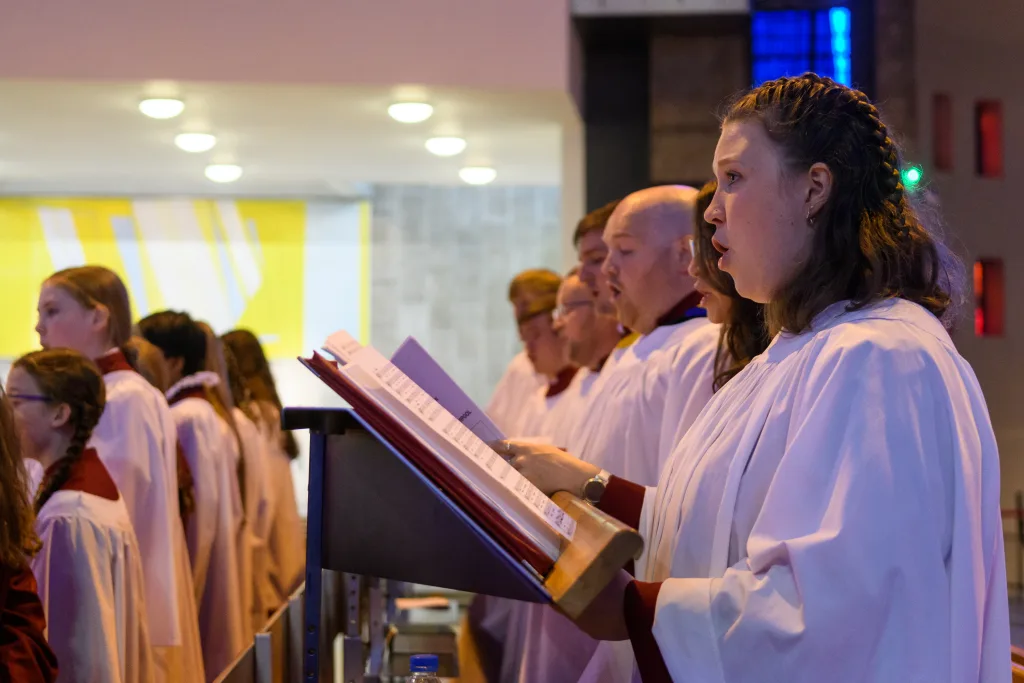 Choir members in white robes with maroon collars stand in a row inside a church, singing from open music folders on a wooden lectern with a young woman in profile at the front.