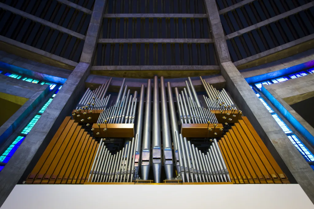 Low-angle view of a large church pipe organ with tall polished metal pipes arranged symmetrically between warm wooden panels, framed by concrete ribs and blue-green stained glass windows.