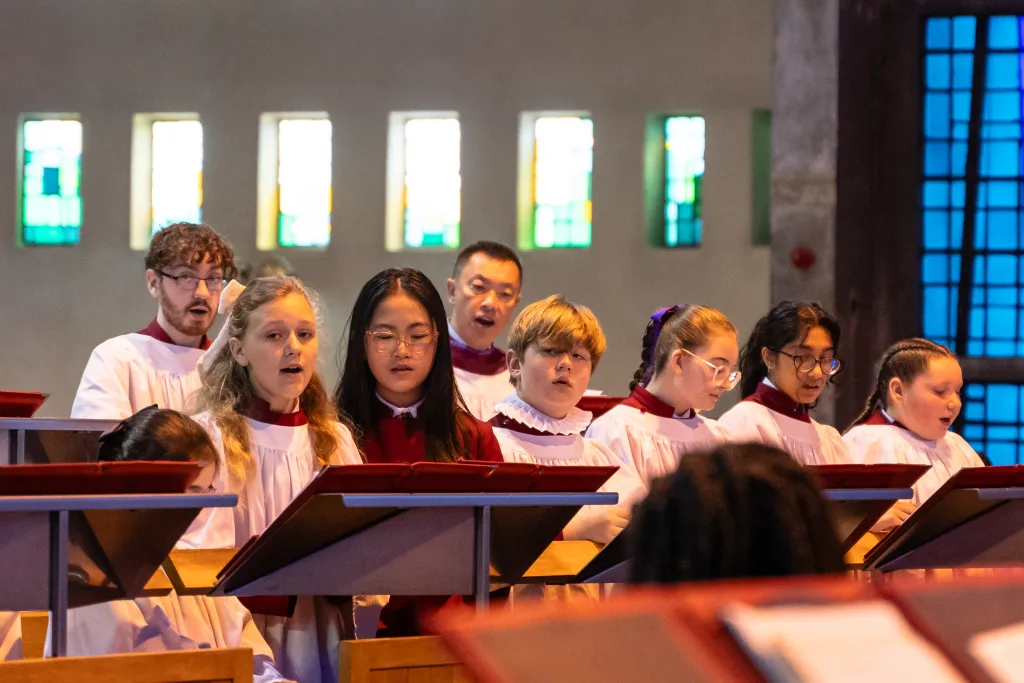 A mixed group of children and an adult choir in white surplices with red collars singing behind wooden music stands, with a row of small coloured stained-glass windows on the church wall behind them.