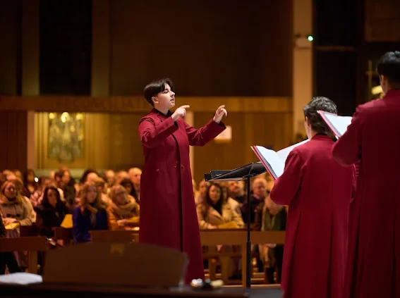 Conductor in a long red robe gestures with raised hands while leading robed choir members singing from sheet music in front of a seated congregation in a warmly lit church interior.