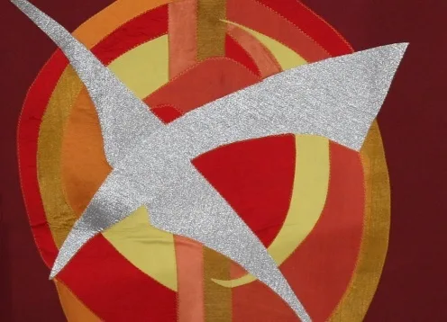 A silver glittering stylised bird-shaped appliqué stretches across concentric red, orange and yellow circular bands on a dark red fabric background.