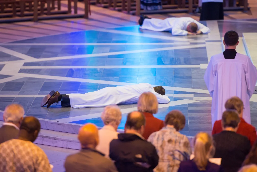 Two people in white liturgical robes lie prostrate on a patterned stone church floor under blue lighting during a religious ceremony, while a robed attendant stands nearby and the congregation watches in the foreground.