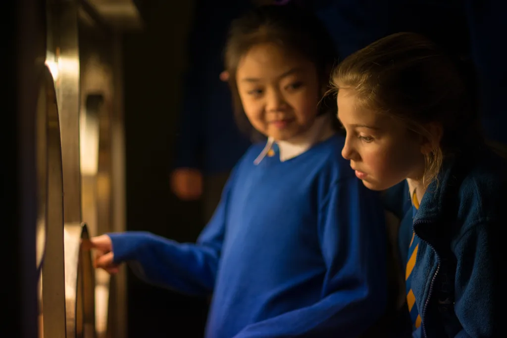Two schoolgirls in blue uniforms lean close to an interactive, softly lit display as one girl points through a circular opening and the other watches intently.