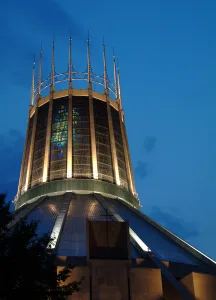 Illuminated circular cathedral lantern with vertical stained-glass panels and tall crown-like spires reaching into a deep blue evening sky.